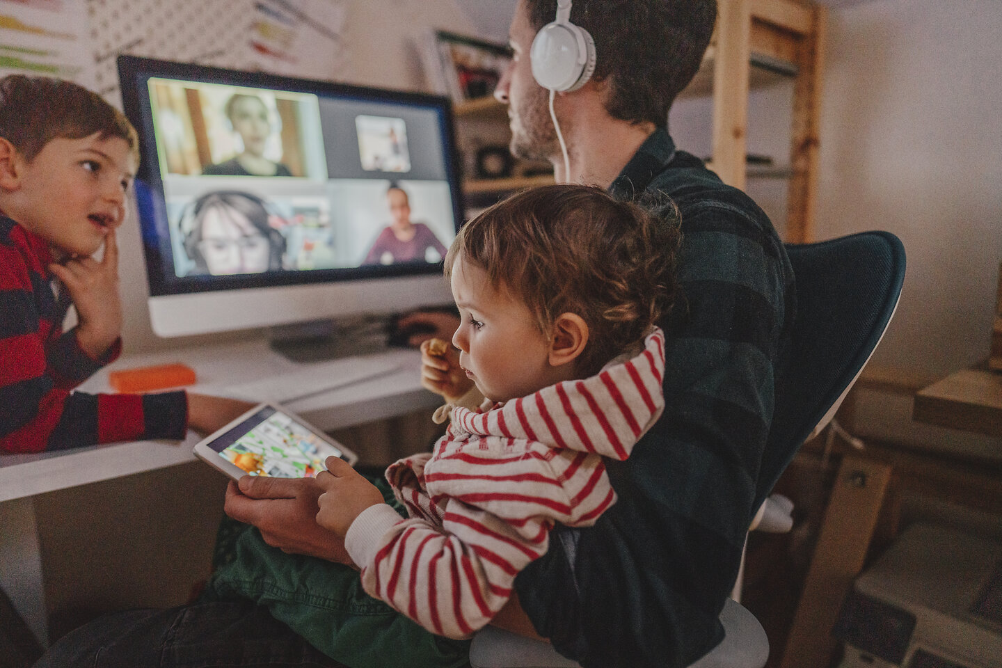 Junger Mann mit Kopfhörern und zwei kleinen Kindern am Schreibtisch im Home Office, der PC überträgt eine Videokonferenz, während ein Kind am Tablet spielt. 