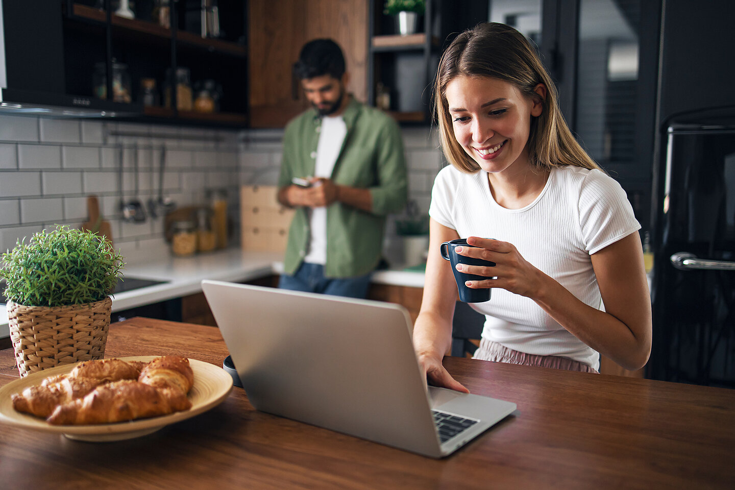 Junges Paar in ihrer Küche, sie sitzt am Laptop. Freund benutzt im Hintergrund sein Mobiltelefon.