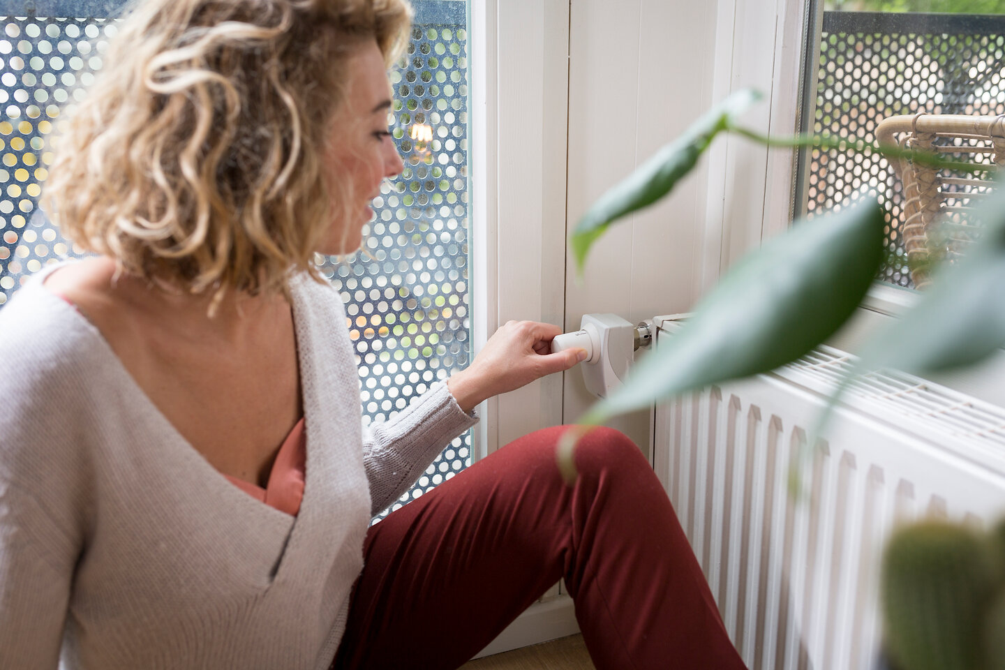 Junge Frau sitzt entspannt auf dem Fußboden in ihrer Wohnung und dreht am Thermostat des Heizkörpers.