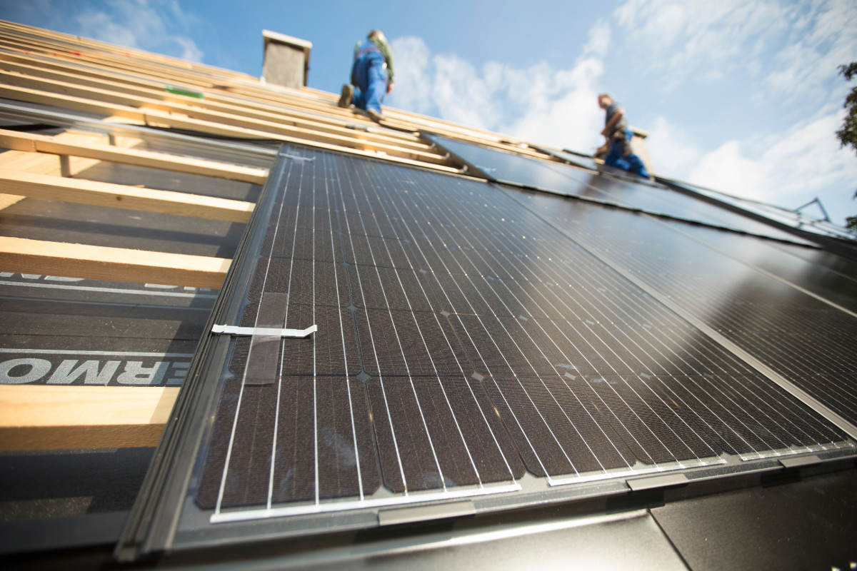 Installation von Photovoltaik auf einem Dach: Module auf Dachlatten von unten fotografiert, oben Schornstein und zwei Handwerker