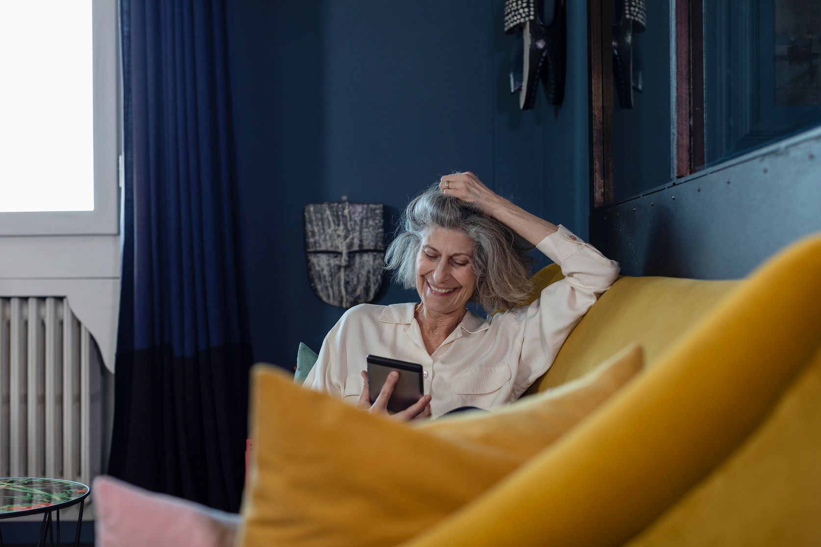 Ältere Frau sitzt auf einem Sofa mit gelben Kissen und schaut auf ihr Smartphone.