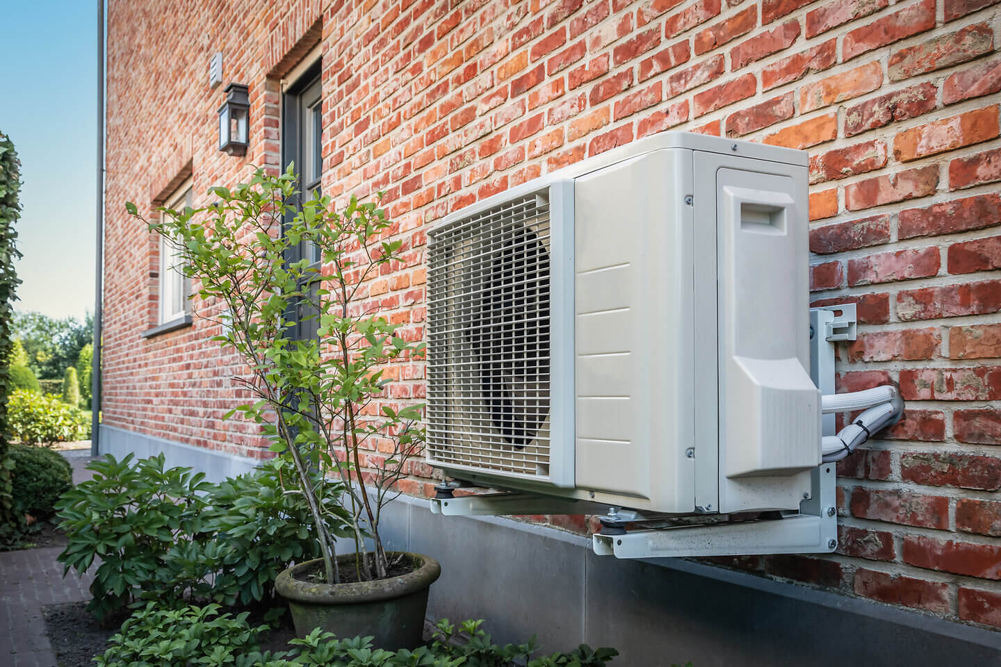 Luftwärmepumpe oder Klimaanlage an der Außenwand eines Hauses mit Backsteinmauer an einem sonnigen Tag mit Pflanzen daneben